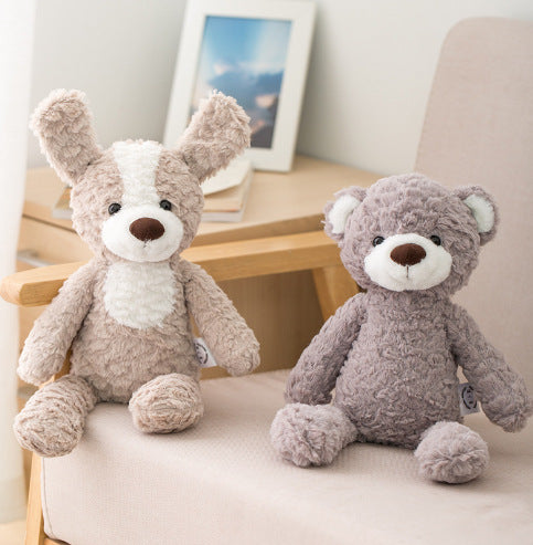 Two plush toys, a beige and white dog with floppy ears and a gray bear, sit on a wooden chair in a cozy room with a framed picture and soft lighting in the background.