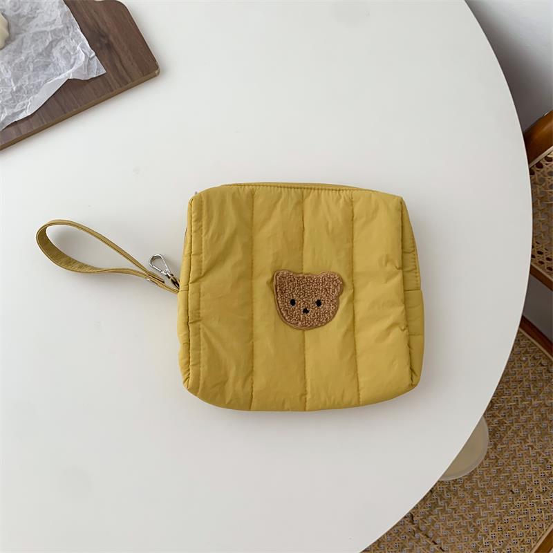 Yellow quilted pouch with a teddy bear patch on a white round table. The pouch has a wrist strap attached. Part of a wooden board with paper is visible beside the pouch.