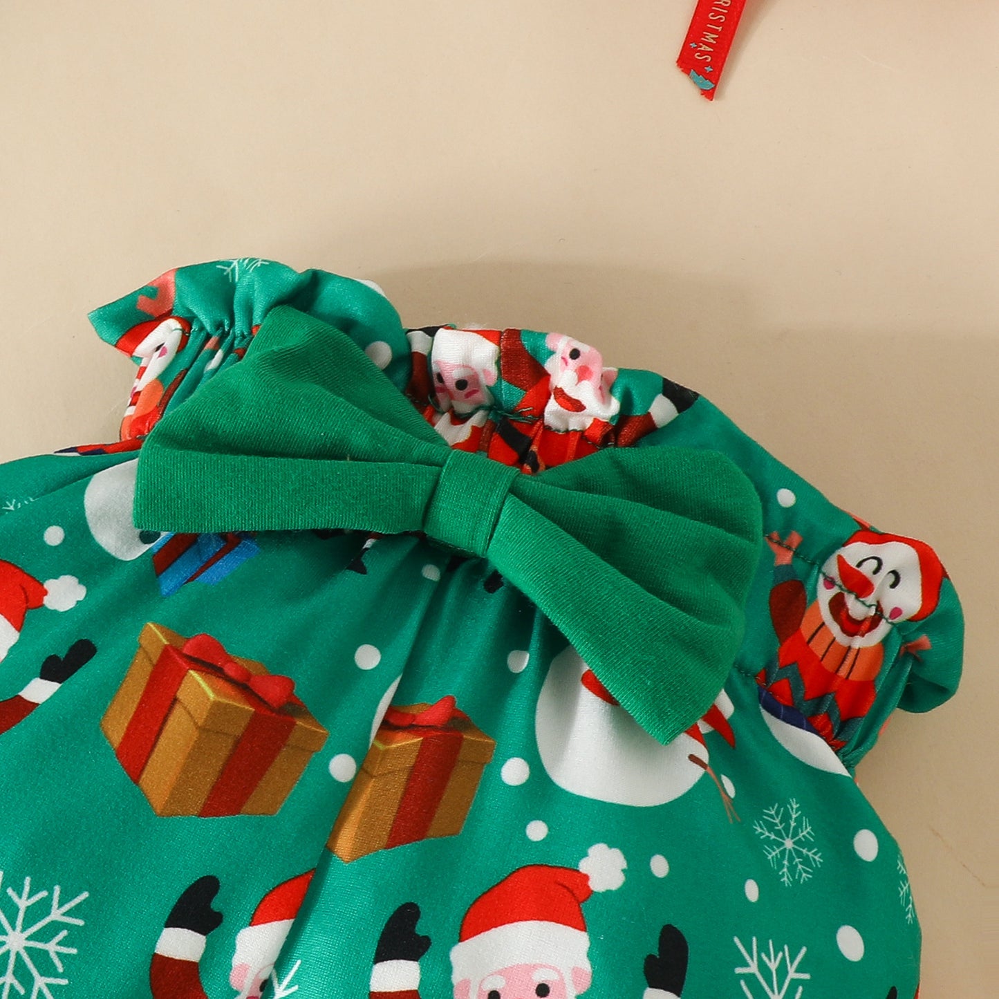 Green Christmas-themed fabric with a big bow, featuring Santas, snowmen, snowflakes, and gifts on a beige background. A small decorative tag is visible at the top.