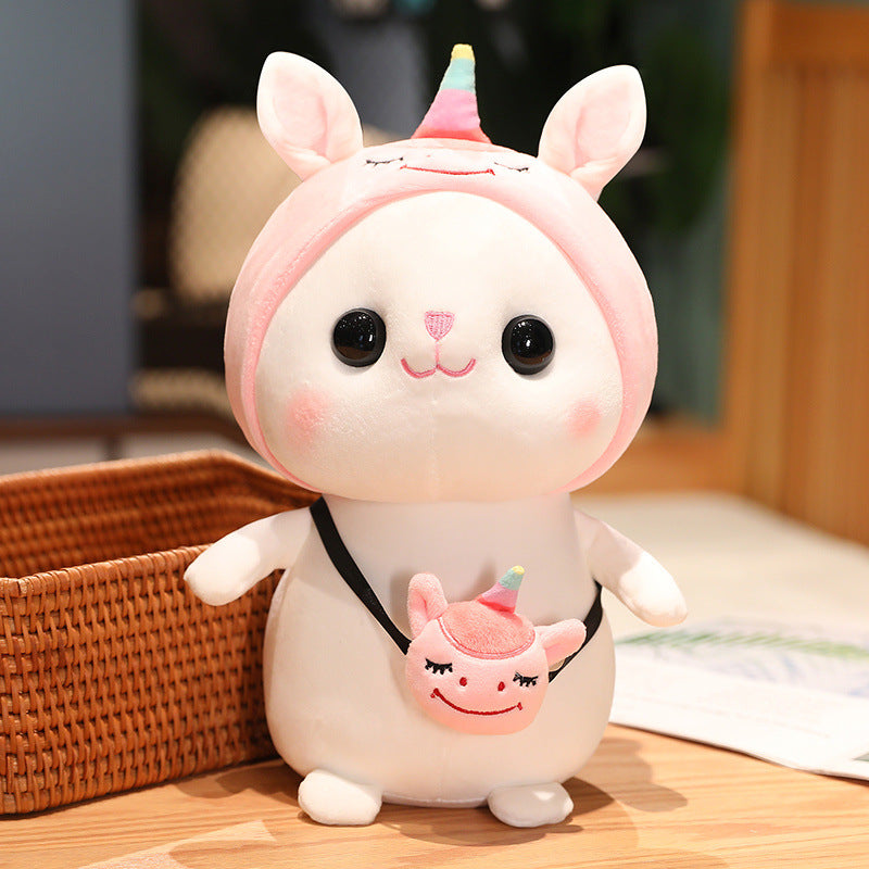 Cute plush toy with large eyes, wearing a pink unicorn hoodie and a small unicorn-themed crossbody bag. It sits beside a woven basket on a wooden surface.