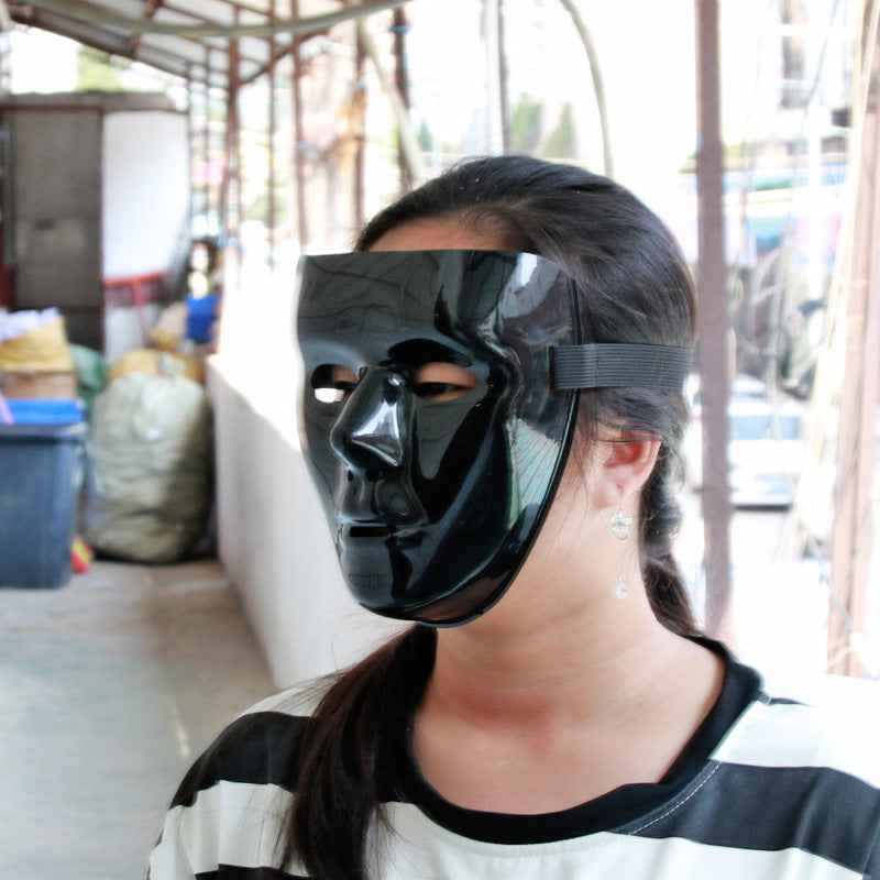 A person with long hair wears a smooth, shiny black mask covering their face. They are standing in a shaded outdoor area near bags and bins. The background is slightly blurred.