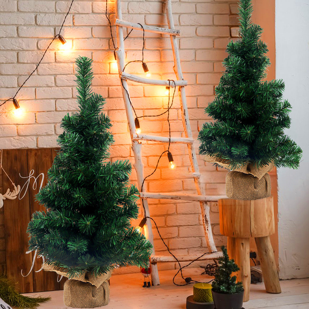 Two small Christmas trees with burlap bases stand against a brick wall, which is adorned with string lights and a rustic ladder, creating a cozy, festive atmosphere.
