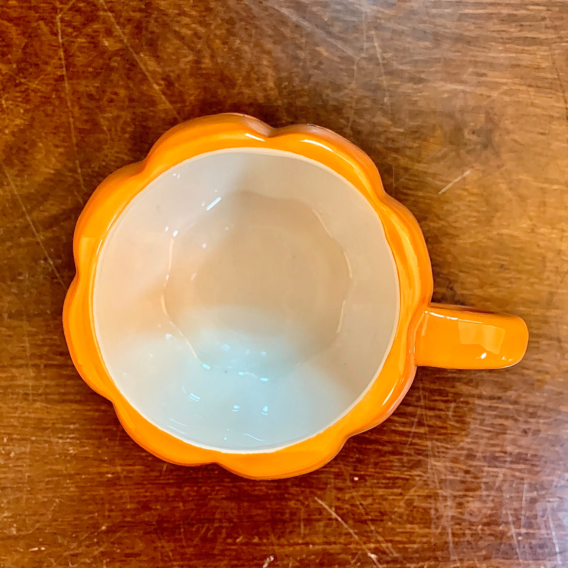 Top view of a ceramic mug with an orange scalloped edge resembling a flower, and a matching handle. The interior is beige. It’s placed on a wooden surface.