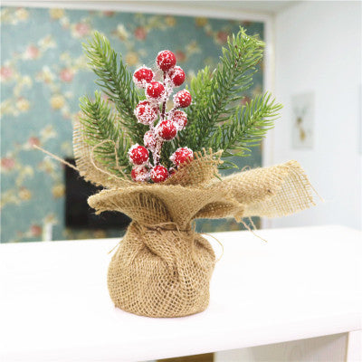 A small decorative plant with frosted red berries and pine branches wrapped in burlap sits on a white surface. The background features floral wallpaper in soft-focus.