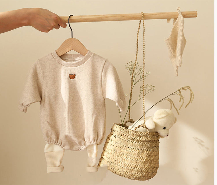 A beige baby outfit with a bear patch hangs on a wooden rod. Below, a woven basket holds a white plush toy and dry flowers. A knitted item also hangs beside the basket.