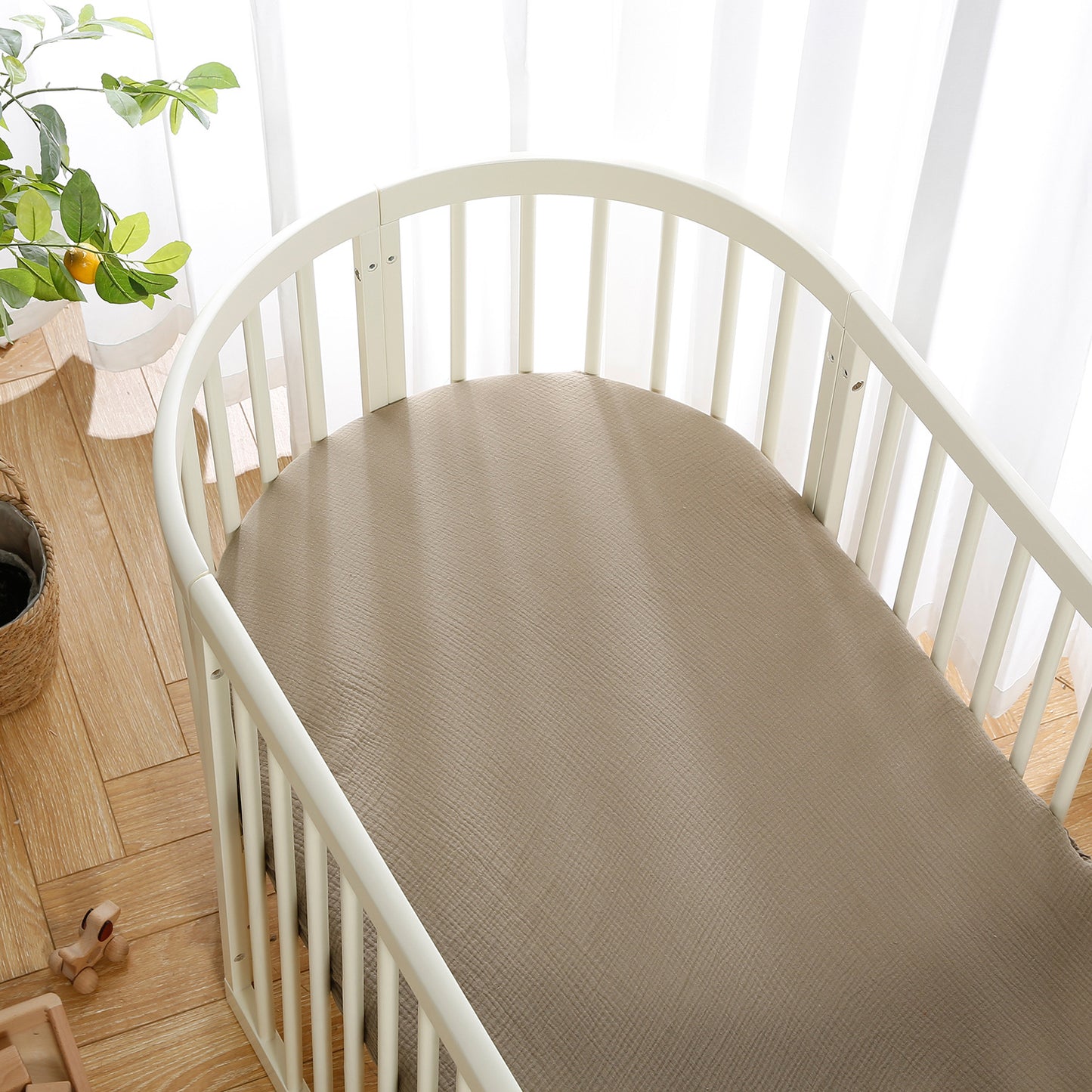 A white oval crib with a beige mattress stands by a window with sheer curtains. A potted plant and a wooden toy are placed on the light wooden floor.
