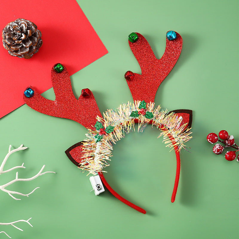 Festive reindeer antler headband with red glitter, colorful bells, and tinsel decoration. Set on green background with red paper, pine cone, white branch, and berries.