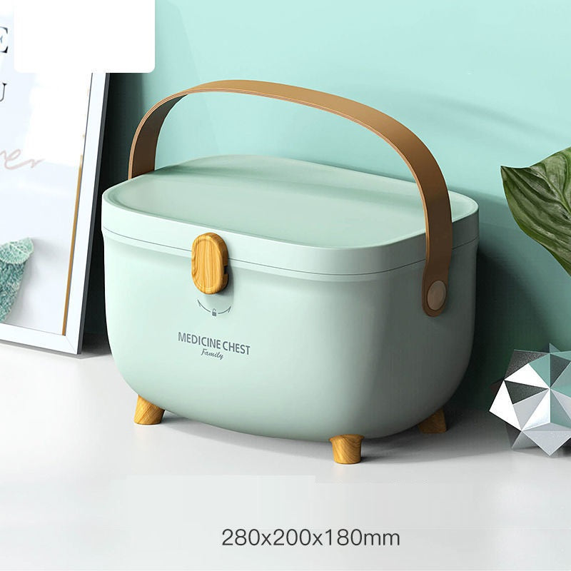 A pastel green medicine chest with a brown handle and wooden latch sits on a white surface. It has small wooden legs and a geometric decoration nearby. Dimensions: 280x200x180mm.