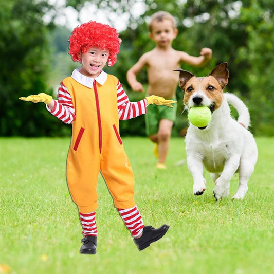 A child in a clown costume poses playfully on grass. A dog runs beside them, holding a tennis ball, while another child follows in the background.