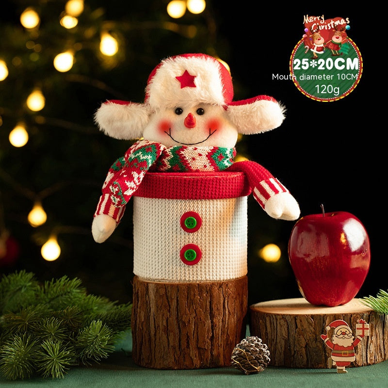Festive snowman decoration with a red and green scarf and hat, sitting atop a log. A red apple and pinecone sit nearby, with blurred Christmas lights in the background.
