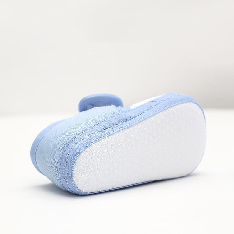 Light blue baby shoe with a soft white dotted sole, viewed from the side. The shoe features fine stitching and a rounded, smooth shape against a plain background.