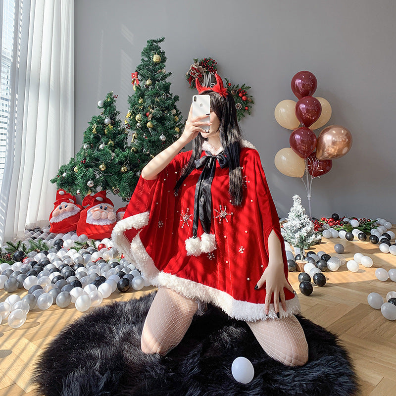 Woman in a red holiday cape kneels on a black rug, taking a selfie. She wears reindeer antlers with decorations around, including a Christmas tree and balloons.
