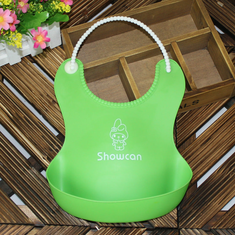 Green silicone bib with a cartoon character design and the word "Showcan" printed. It has a beaded strap and is placed on a wooden surface near a box and flowers.