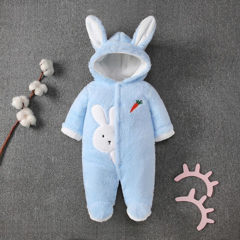 Blue infant onesie with bunny ears and a rabbit design holding a carrot on the chest. A cotton branch is on the left and pink decorative lashes on the right, resting on a gray background.