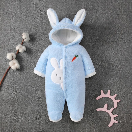 Blue infant onesie with bunny ears and a rabbit design holding a carrot on the chest. A cotton branch is on the left and pink decorative lashes on the right, resting on a gray background.