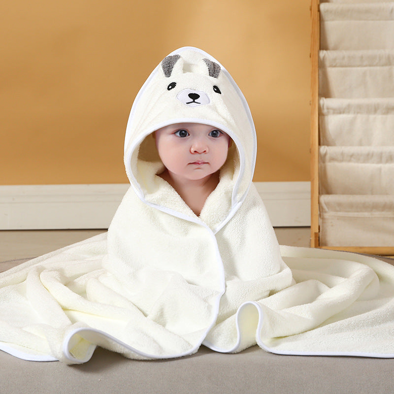 A baby is wrapped in a soft, white hooded towel with bear ears and a face design, sitting on a light-colored surface. The background includes a beige wall and a fabric organizer.