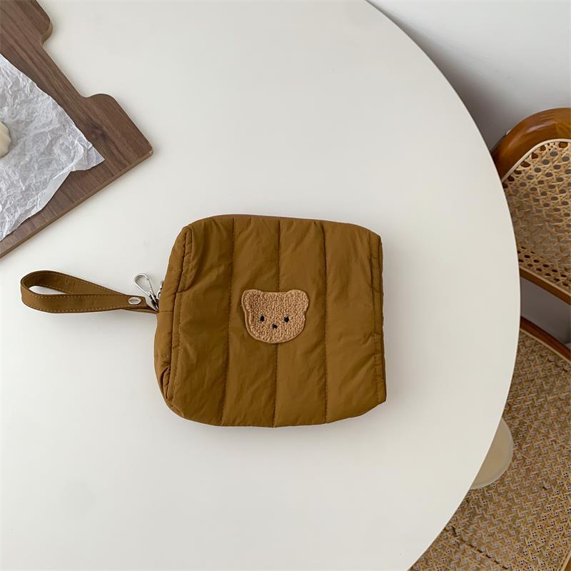 Brown quilted pouch with a bear emblem on a round white table, next to a wooden board with parchment paper. A wicker chair is partially visible to the side.