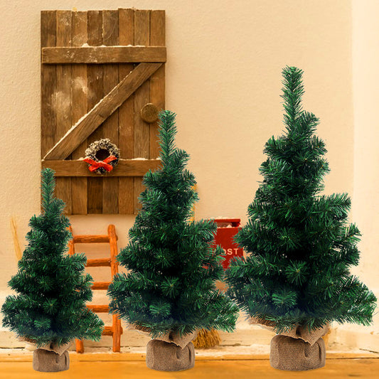 Three small artificial Christmas trees in burlap sacks stand before a rustic wooden door adorned with a tiny wreath. A red mailbox and orange ladder sit in the background.