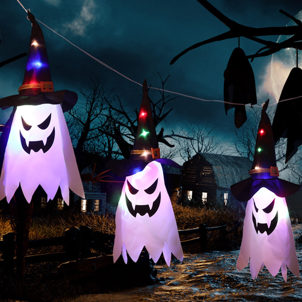 Hanging ghost decorations with witch hats and colorful lights illuminate a spooky outdoor scene with a moonlit sky, barren trees, and a rustic house in the background.
