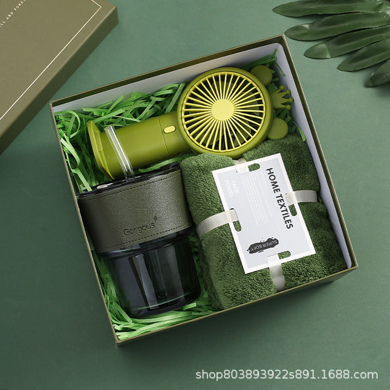 Gift box containing a green portable fan, a dark green insulated cup, and a soft green towel labeled "Home Textiles." The items rest on green shredded paper.