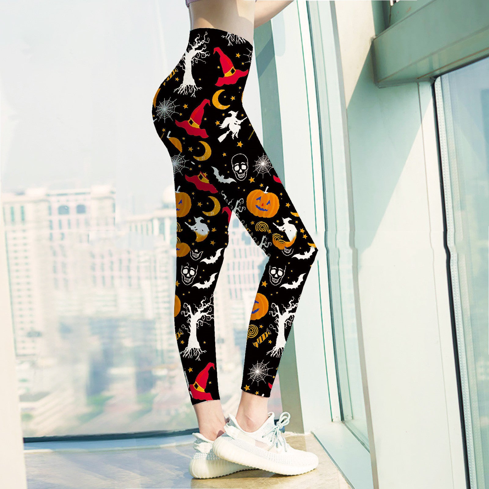 Woman wearing Halloween-themed leggings with designs of pumpkins, witches' hats, skulls, and trees. She stands by a window with a cityscape in the background.