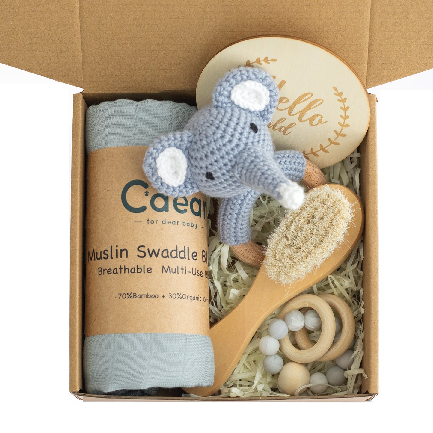 Gift box with a muslin swaddle, crocheted elephant toy, wooden baby brush, engraved wooden plaque, and baby teething rings, set on shredded paper bedding.