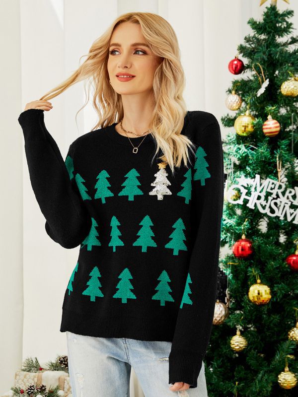 A woman with blonde hair wearing a black sweater featuring green tree patterns and a single silver tree. She's standing by a decorated Christmas tree with a "Merry Christmas" sign.
