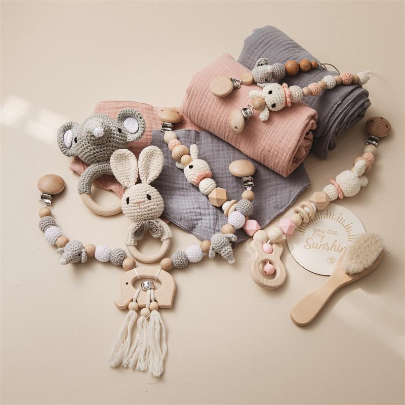 Crochet baby toys and accessories on soft pink and gray blankets include a bunny, elephant, and bear-themed teethers with wooden beads, next to a brush and "You are my sunshine" sign.