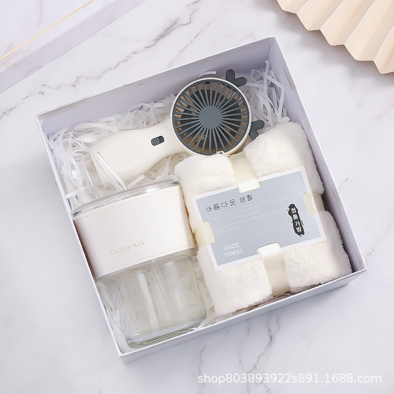 A neatly arranged gift box containing a portable fan, a white face towel, and a glass cup with a cover, set on a white marble background.