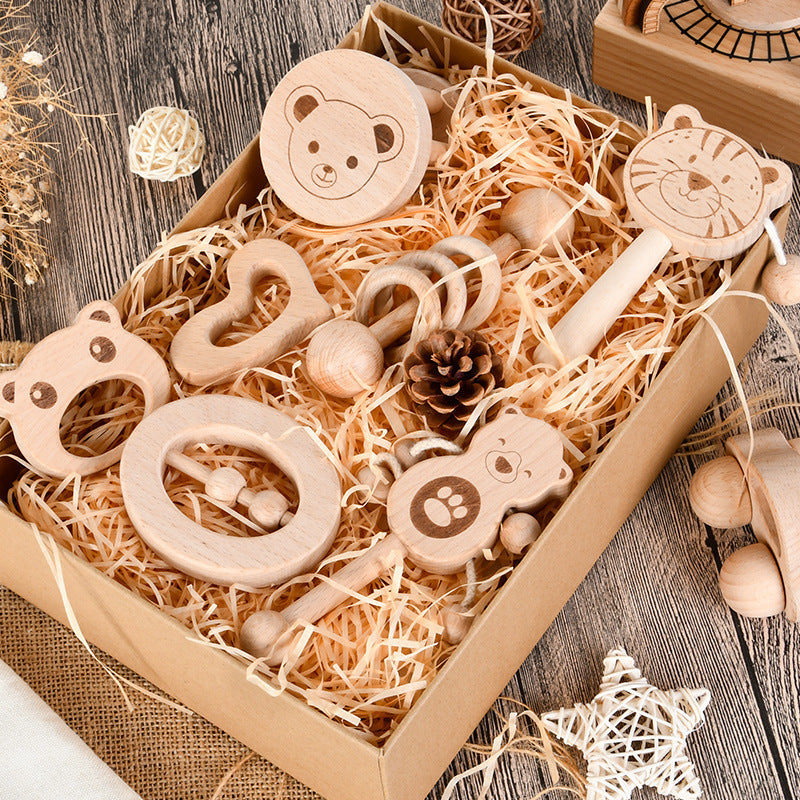 A box of wooden baby toys on a straw bed includes bear and tiger face rattles, a heart-shaped teether, and a pinecone, set on a wooden surface with decorative elements.