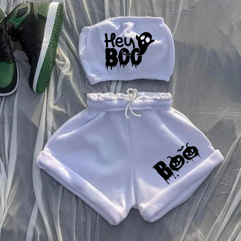 Two-piece outfit includes a white crop top with "Hey Boo" and a ghost graphic, paired with matching shorts featuring "Boo" and pumpkin faces. The set lies on a sheer fabric next to green and black sneakers.