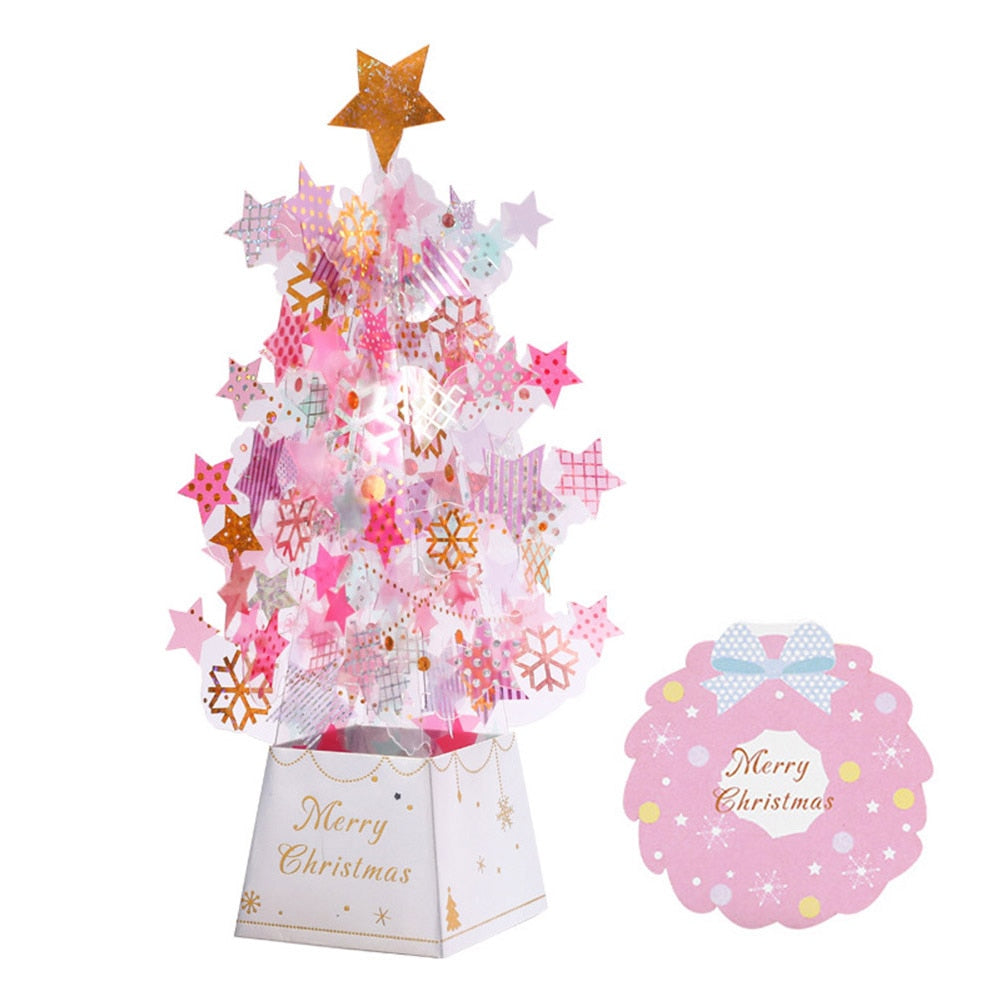 Pink and gold Christmas tree with a star topper, adorned with stars and snowflakes, on a white "Merry Christmas" base. Beside it is a pink card with a bow and festive message.