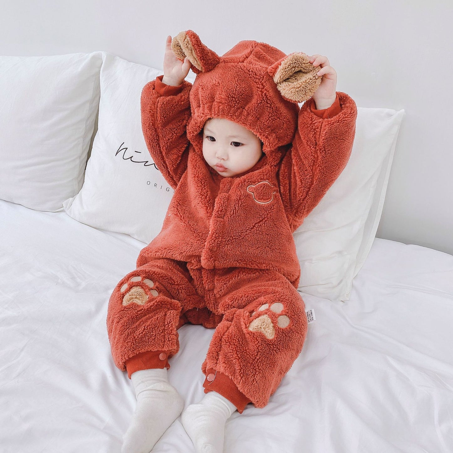 A baby in a cozy red bear-themed outfit with ear and paw details sits on a bed. The baby is lifting the hood's ears, with white pillows in the background.