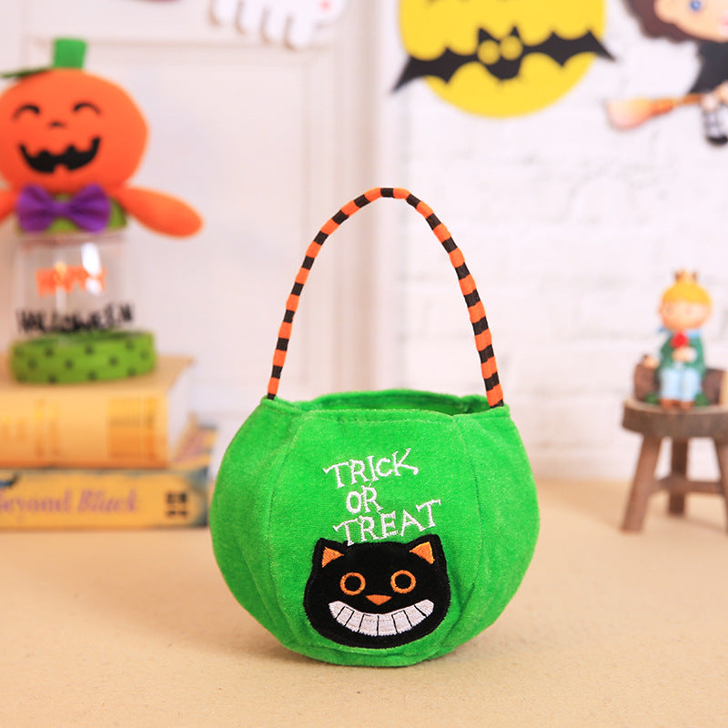 Green trick-or-treat bag with black cat design and "TRICK OR TREAT" text, featuring an orange and black striped handle. Halloween decorations are blurred in the background.