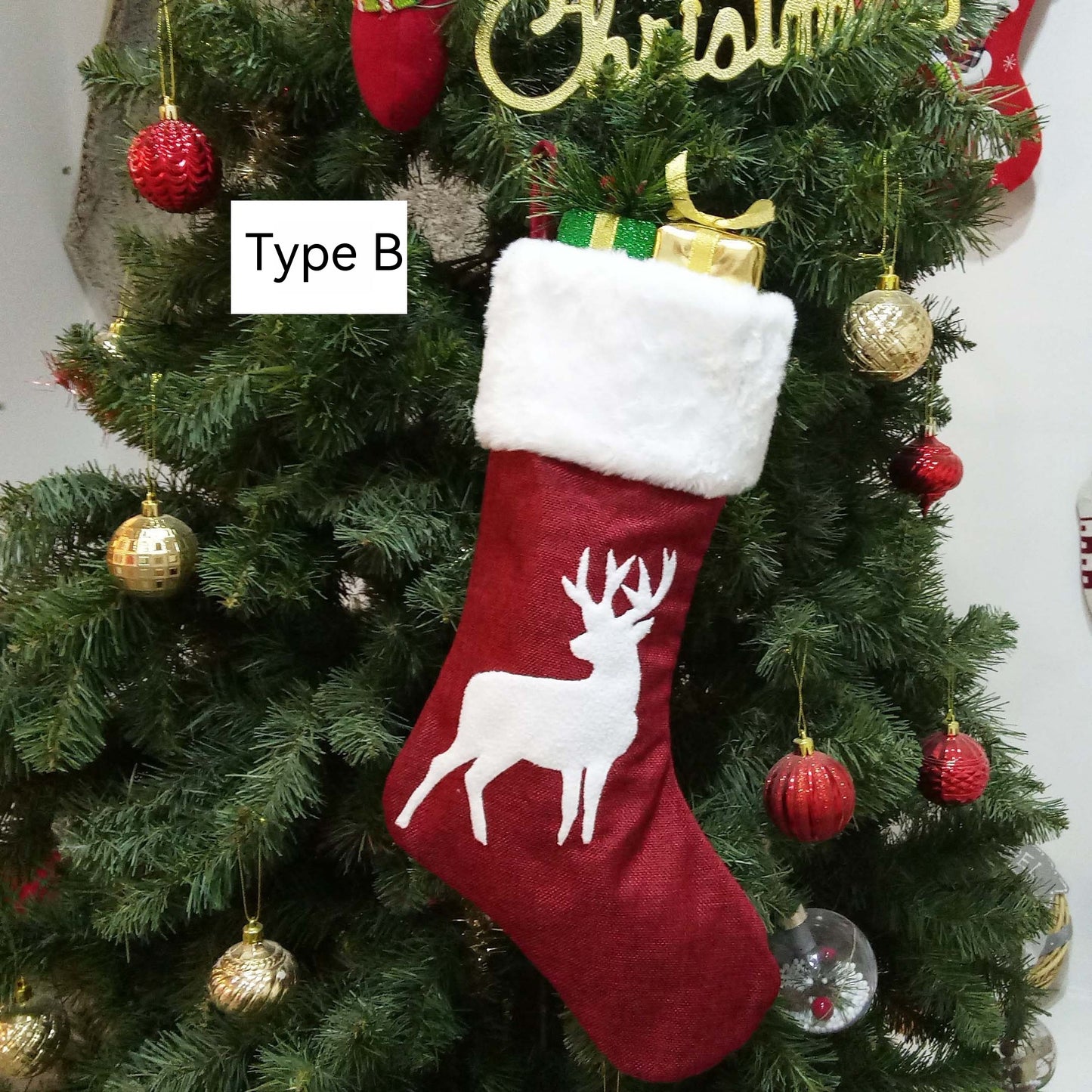 A red Christmas stocking with a white reindeer design hangs on a decorated tree with gold and red baubles. The stocking is filled with a green and gold present.