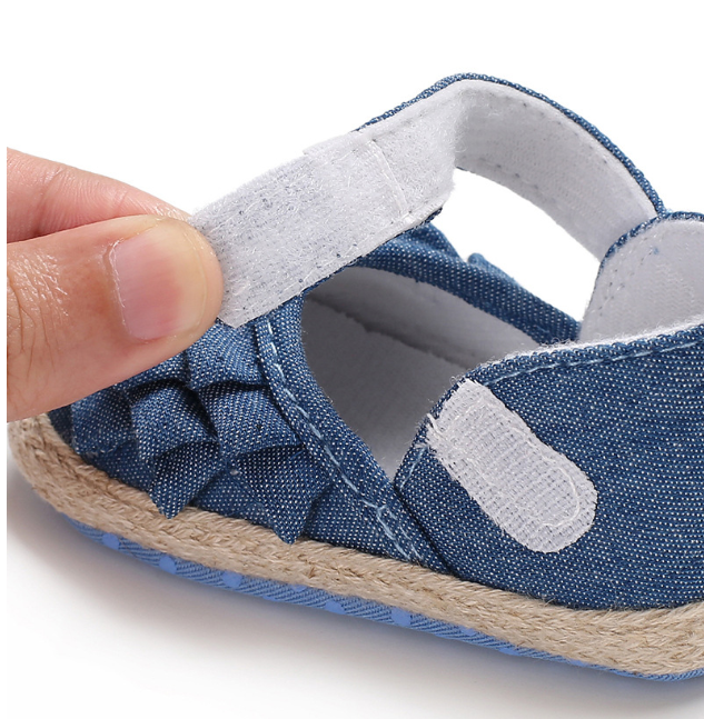Close-up of a hand holding the Velcro strap of a blue denim baby shoe with a decorative bow and textured sole, showing ease of fastening.