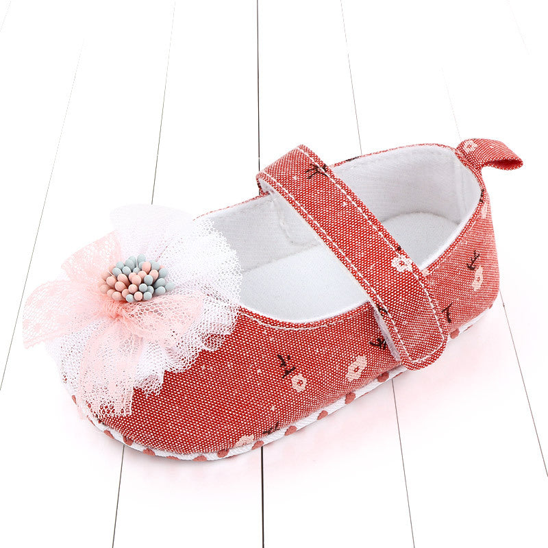 Red baby shoe with white floral pattern, featuring a lace flower embellishment with pastel beads. It has a strap and is placed on a white wooden floor.