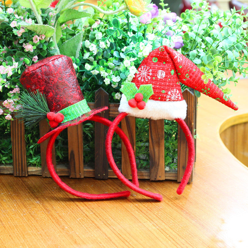 Two festive headbands rest on a wooden surface. One features a red top hat with green accents, while the other displays a red Santa hat with white trim and holly, set against greenery.