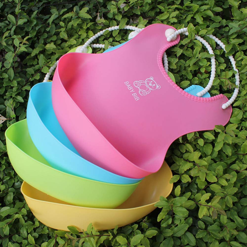 Four colorful silicone baby bibs in pink, blue, green, and yellow are stacked on green foliage. Each bib features a beaded adjustable neck strap and a deep pocket.