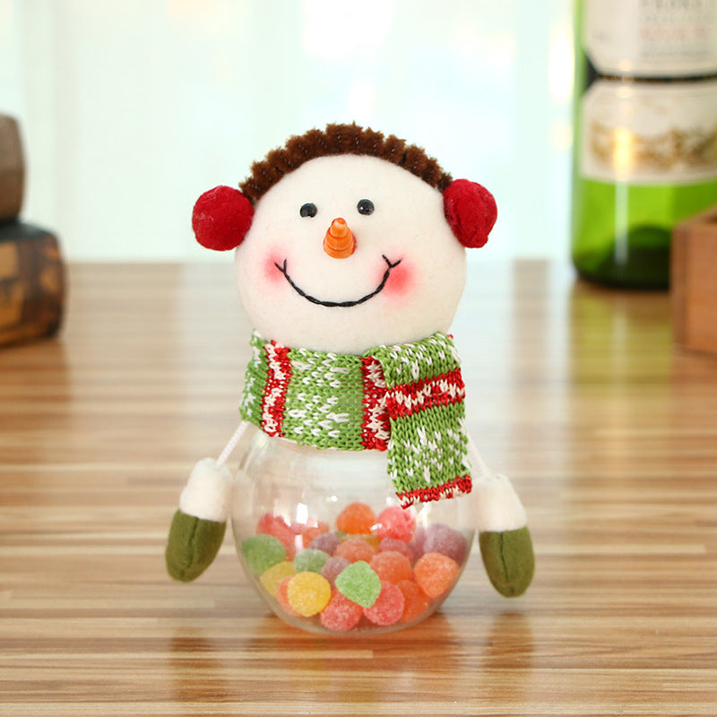 A snowman figurine with red earmuffs and a green and red scarf, smiling on a wooden surface. Its clear lower half contains colorful candy. Background shows blurred bottles.