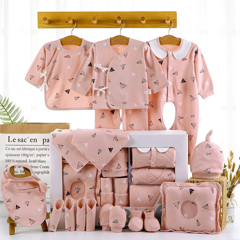 Pink baby clothing set with paper airplane pattern, displayed on hangers and a shelf. Includes onesies, pants, bib, booties, hat, and blanket. A plant and bag are in the background.