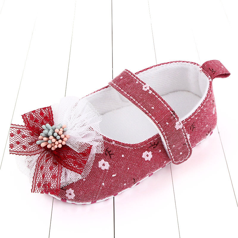 Red baby shoe with white floral patterns, featuring a lace bow and small pink and gray embellishments on the toe. The shoe has a strap and is placed on a white wooden surface.
