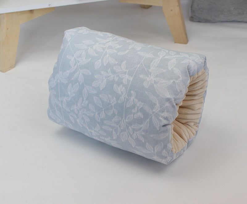 A cylindrical pillow covered in light blue fabric with a white leaf pattern, neatly rolled and placed on a white surface. The edges reveal a beige interior.