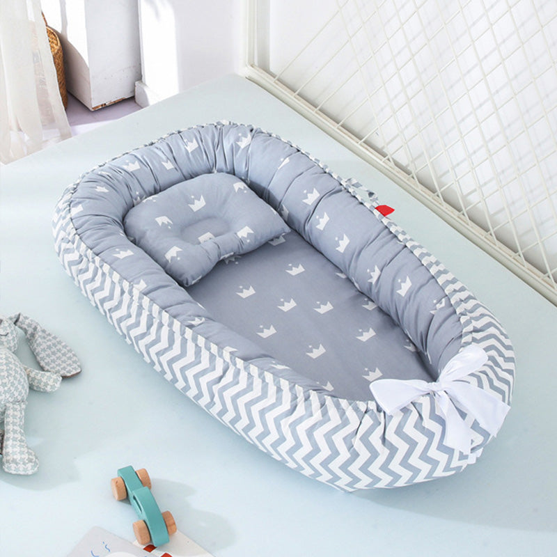 Gray baby nest with white crown prints and zigzag edge pattern, adorned with a white bow. Nearby are a teal wooden toy car and a stuffed bunny on a light blue surface.
