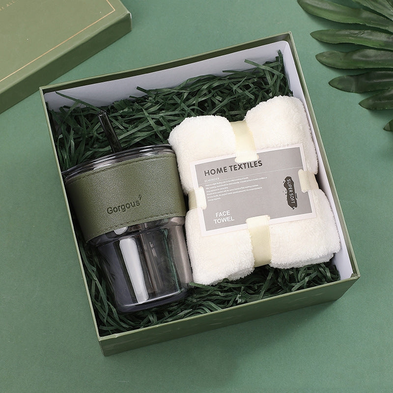 Gift box with a green-lidded tumbler and a white face towel labeled "Home Textiles" on a green shredded paper base, set against a green background with leaves.