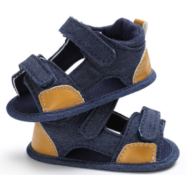Two denim baby sandals with velcro straps stacked together. They feature tan leather patches on the toe and heel, and have a dark blue inner lining.