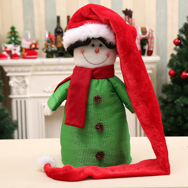 Festive snowman decoration wearing a long red Santa hat and scarf, dressed in a green outfit with button details, set against a Christmas-themed room with a fireplace and tree.