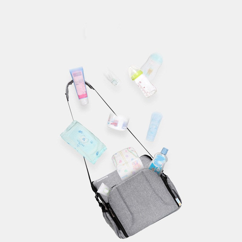 Gray diaper bag with numerous baby essentials spilling out, including a feeding bottle, wipes, lotion, diapers, and creams, set against a plain white background.