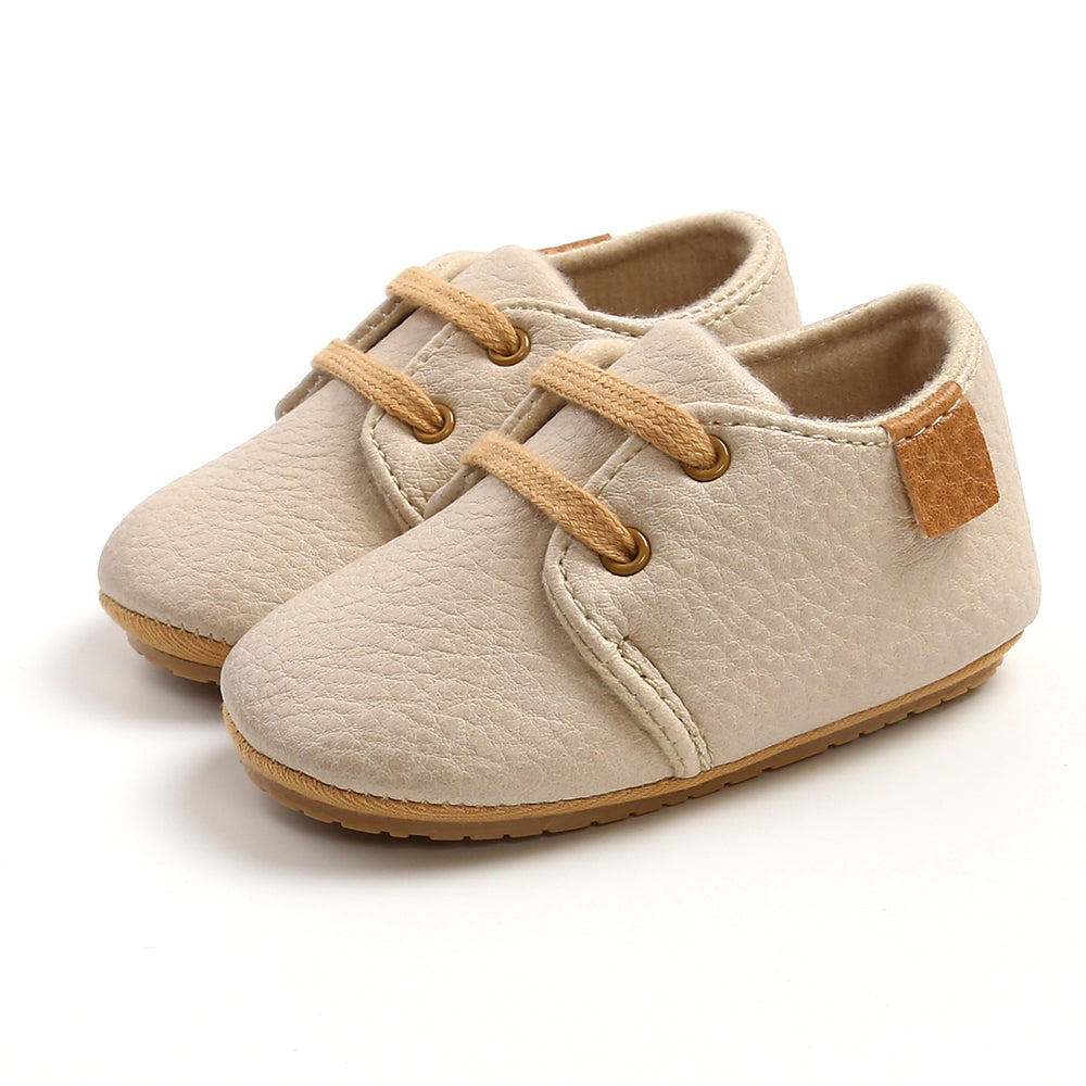 Beige baby shoes with textured soft leather, light brown laces, and a small brown patch on the side, designed for comfort and style, placed on a white background.