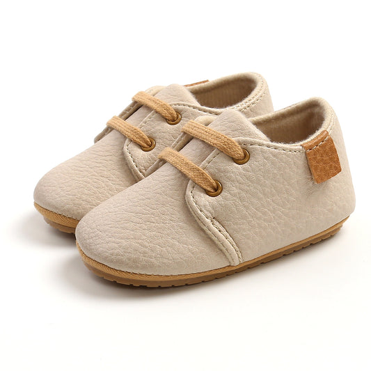 Beige baby shoes with textured soft leather, light brown laces, and a small brown patch on the side, designed for comfort and style, placed on a white background.